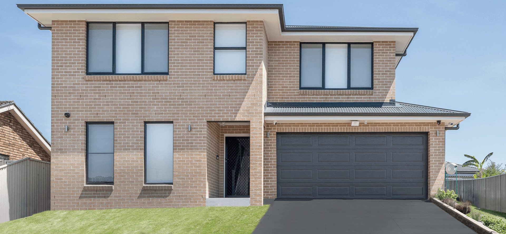 A modern two-story brick house with large windows, a black double garage door, and a small front lawn, set under a clear blue sky.