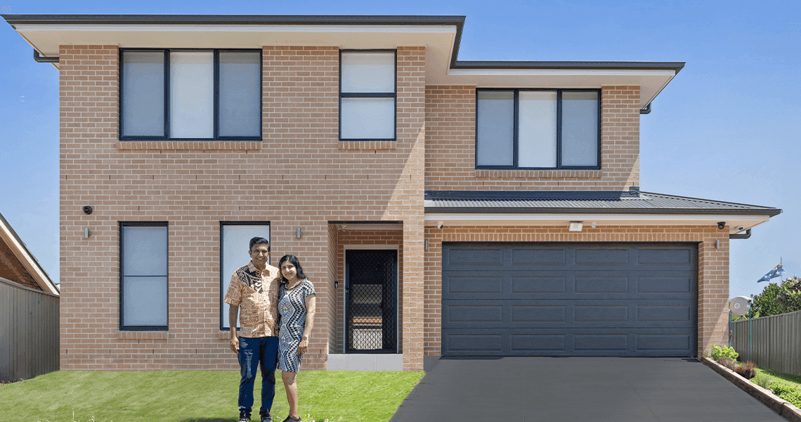 A couple stands on the lawn in front of a modern two-story brick house with large windows and a double garage on a sunny day.