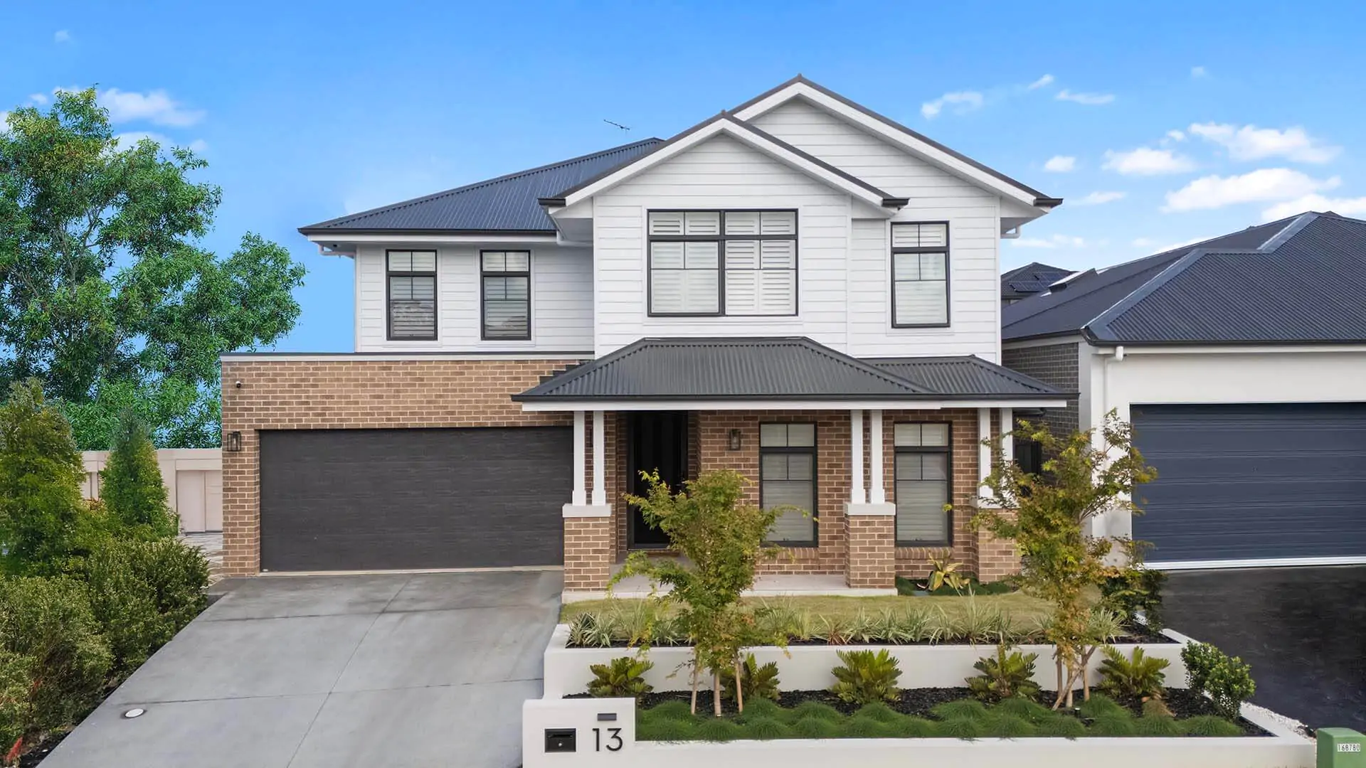 A modern two-story house with brick and white siding exterior, black roof, double garage, front porch, small garden, and driveway, set against a blue sky.