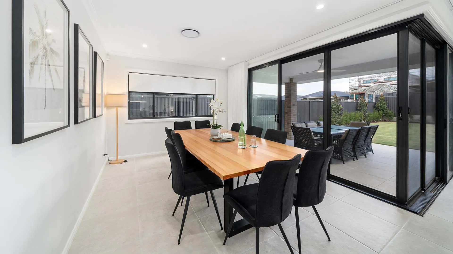 Modern dining room with a wooden table and eight black chairs, large glass doors open to a patio and garden. Minimalist decor with wall art, a floor lamp, and neutral tones throughout the space.