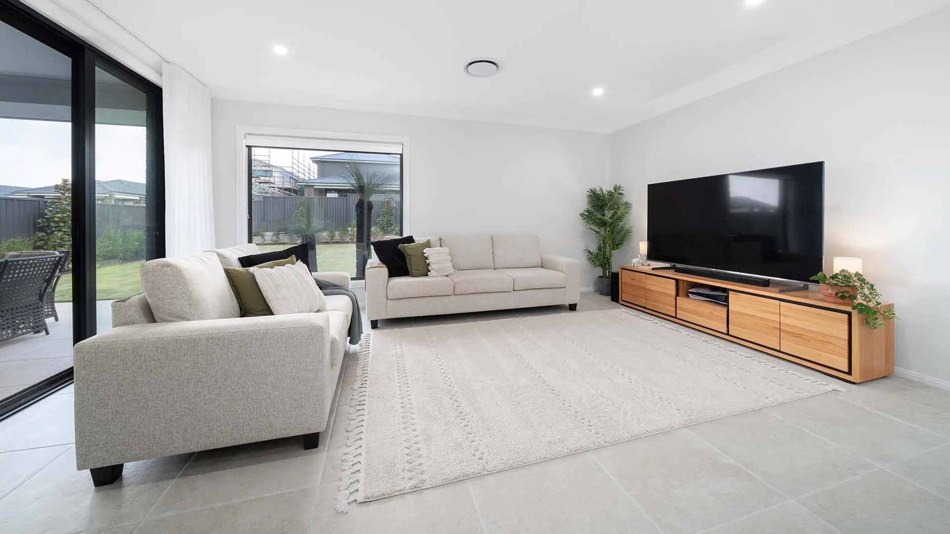 Modern living room with two light-colored sofas, a large flat-screen TV on a wooden stand, indoor plants, neutral decor, and large windows overlooking a backyard with green grass. The room is bright and spacious.