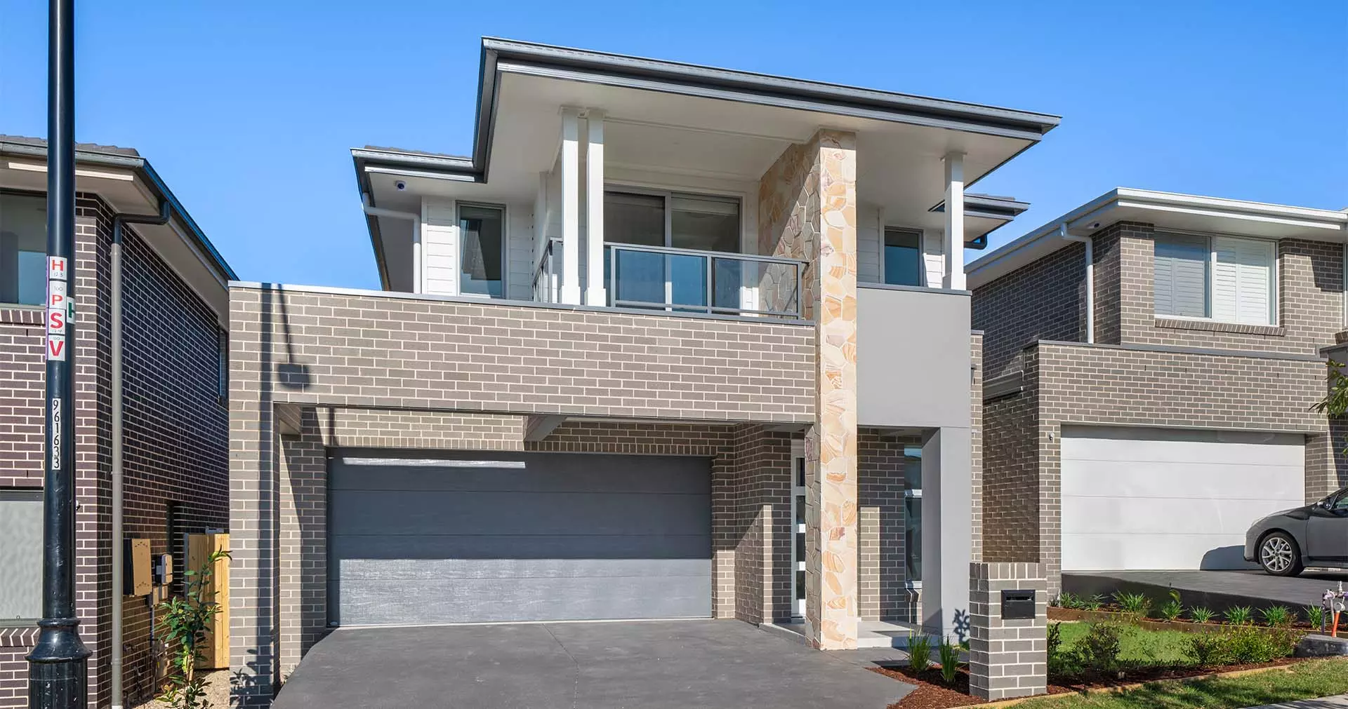 Modern two-story house with gray brick exterior, double garage, a stone accent pillar, balcony with glass railing, and well-maintained front yard under a clear blue sky.