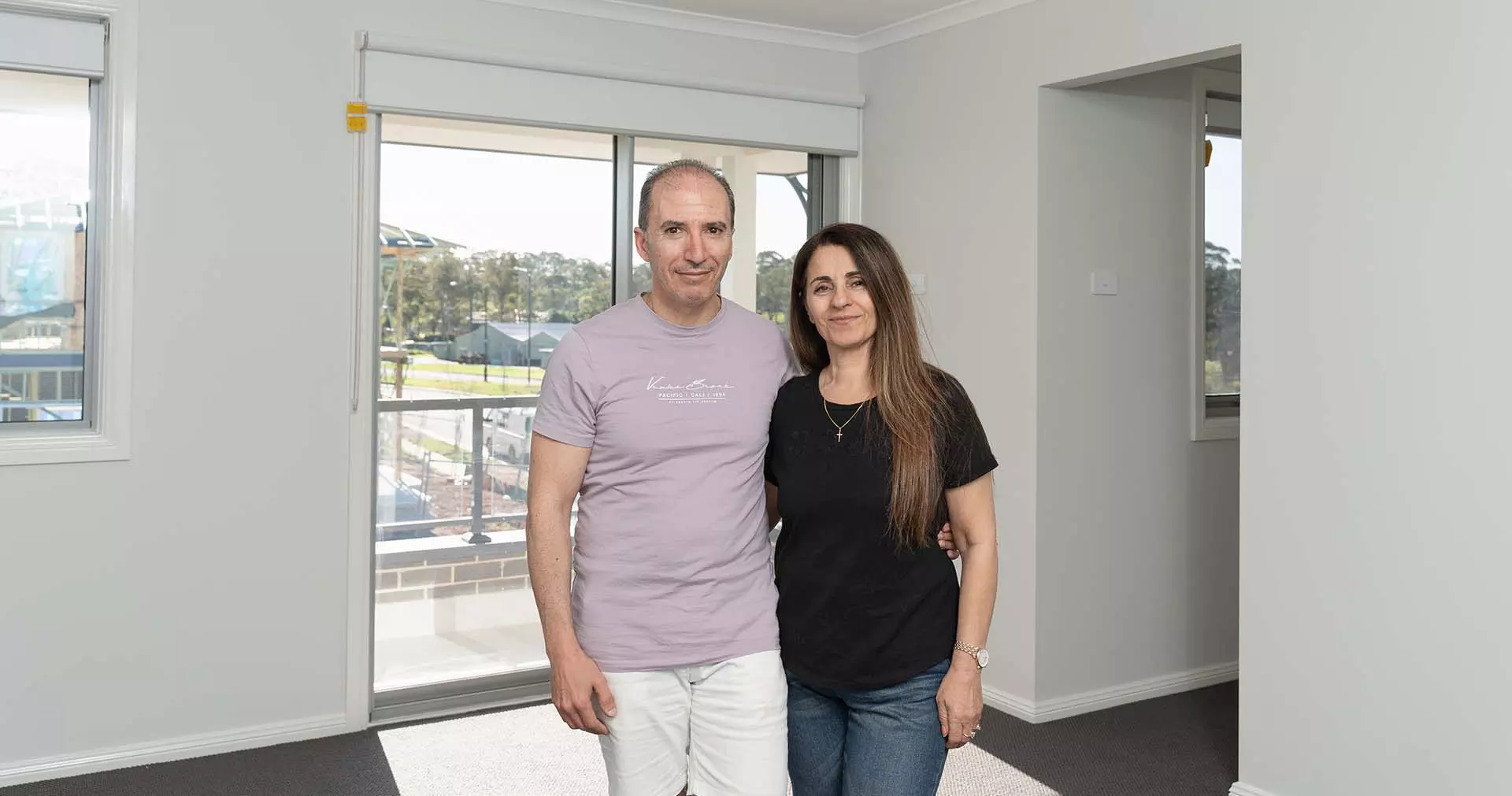 A man and woman stand together in a bright, empty room with large windows and a sliding glass door, smiling at the camera.