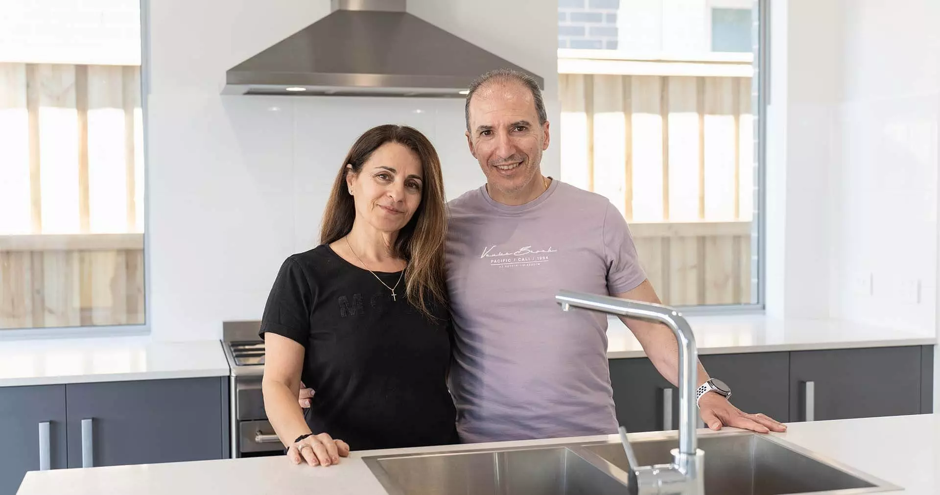 A man and a woman stand together in a modern kitchen, smiling at the camera. The kitchen features a stainless steel sink, white countertops, and dark cabinets. Natural light comes through two large windows in the background.