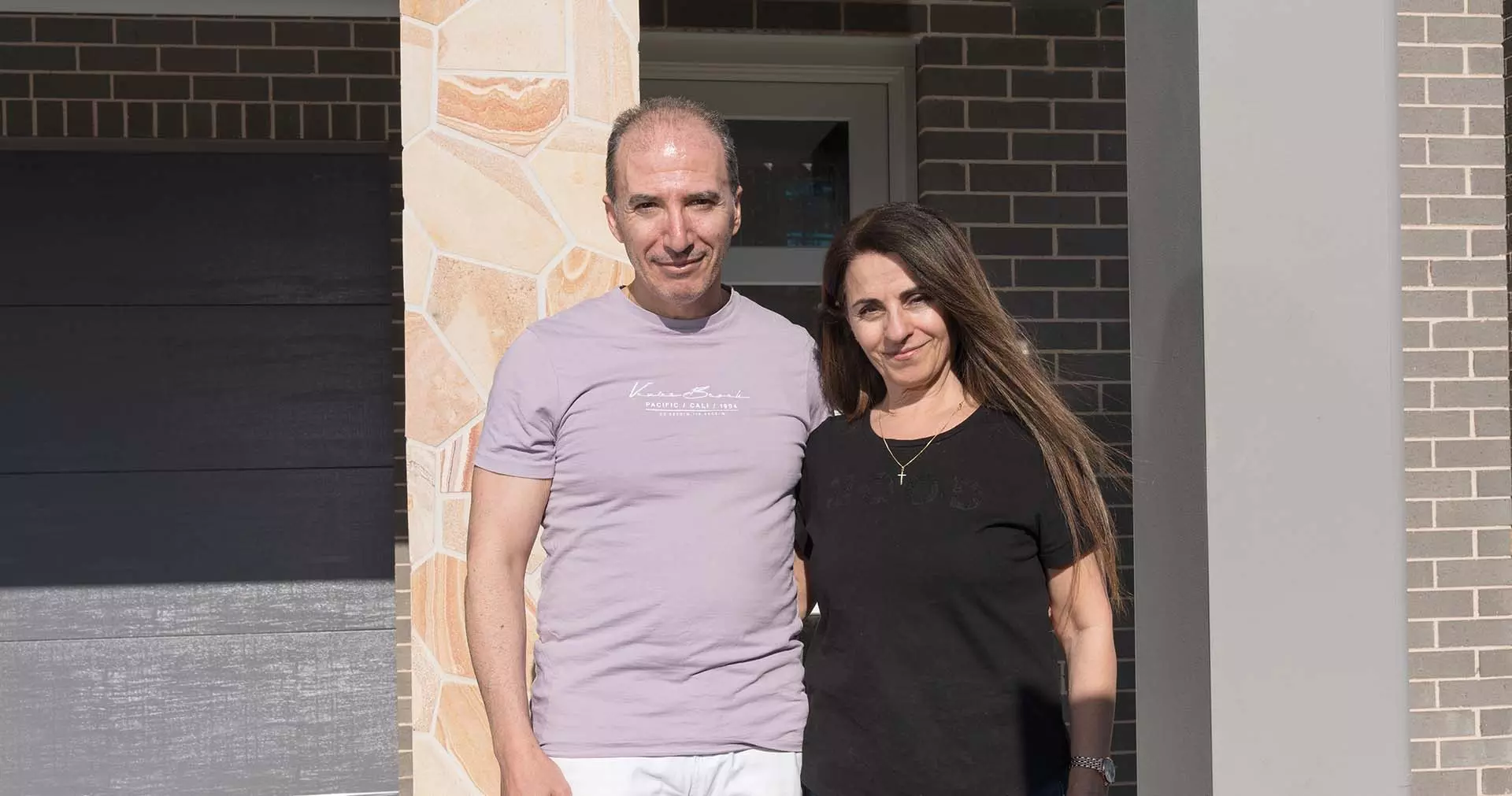A man and a woman stand close together, smiling at the camera, in front of a modern brick and stone building on a sunny day.