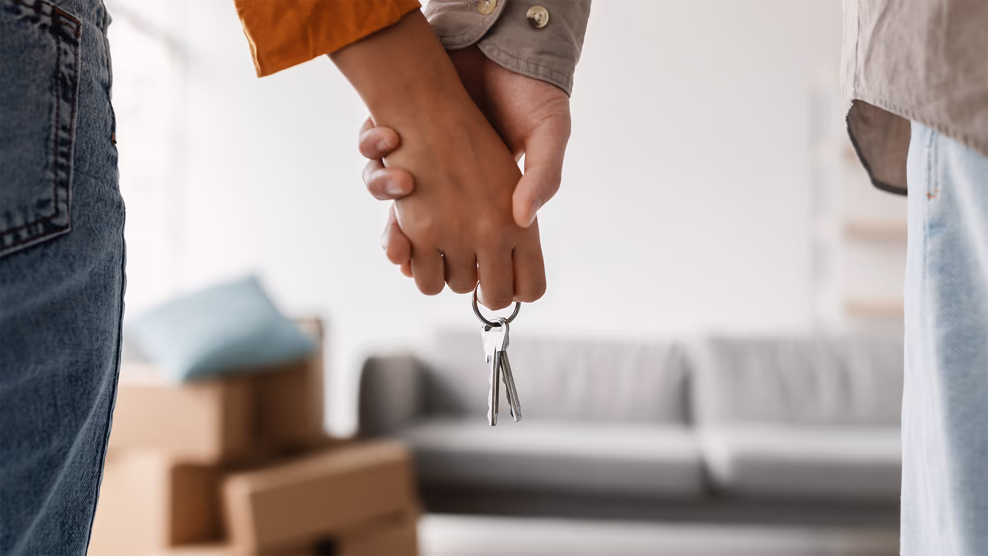 A close-up of two people holding hands with a set of keys, standing in front of cardboard boxes, suggesting moving into a new home.